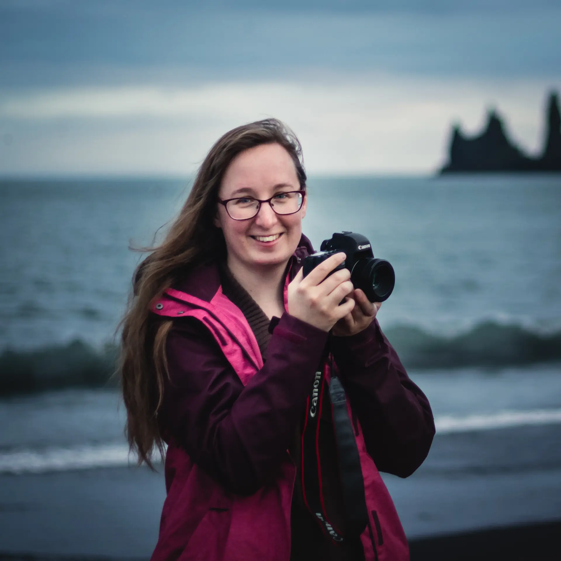 Landscape photographer Nina Lozej, smiling as she holds a DSLR camera on a moody beach.