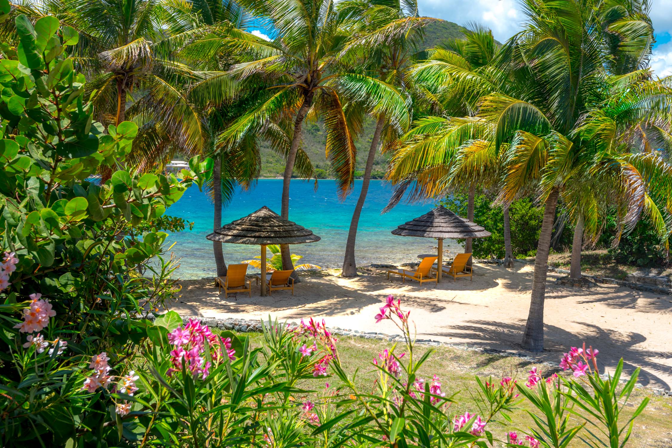 Two thatched-roof tiki huts with lounge chairs sit on a white sand beach under palm trees and turquoise water.