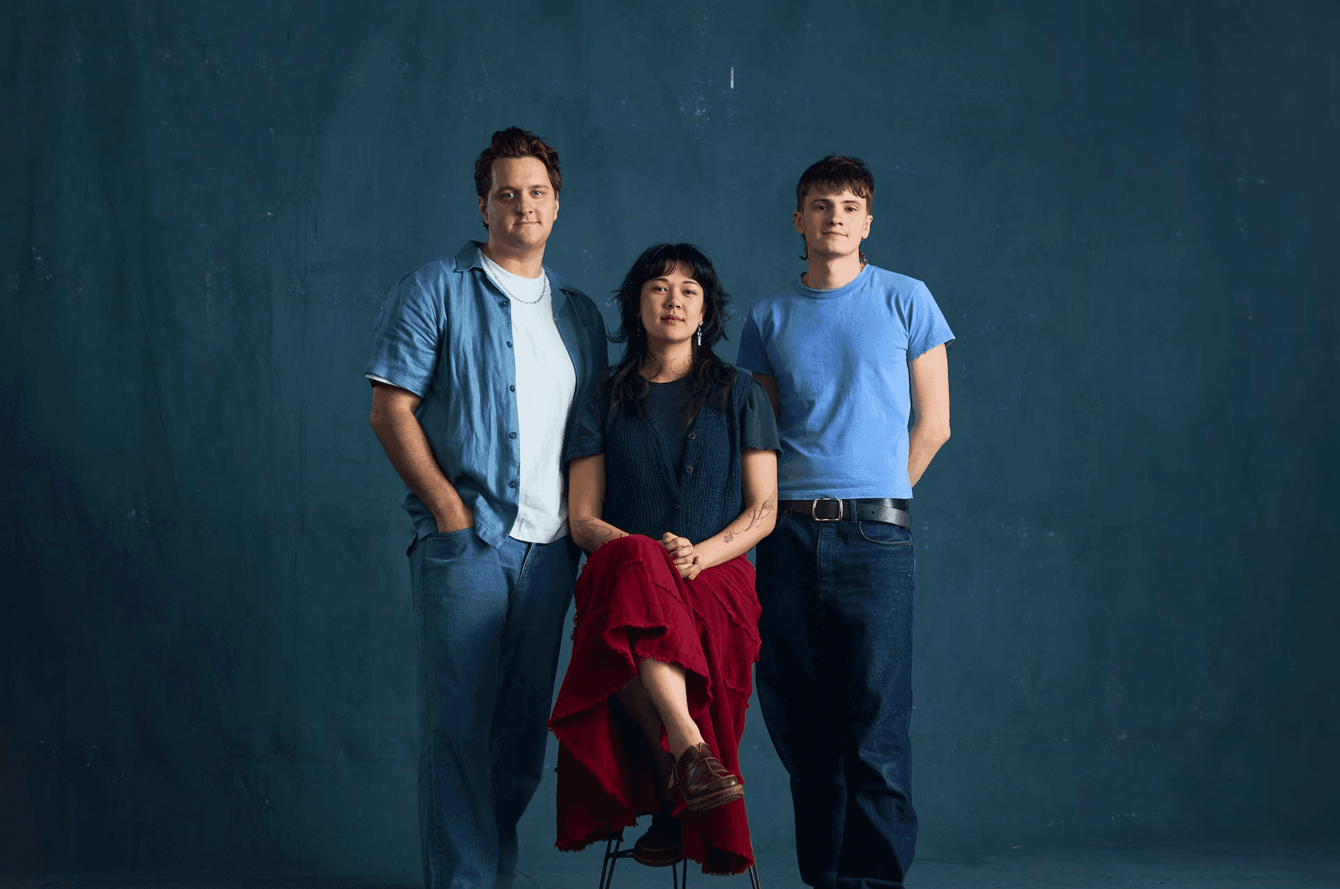 A group portrait of three young adult theatre artists standing and sitting against a dark teal backdrop. A person wearing a navy top and red skirt is seated on a stool in the centre, while two others stand on either side, dressed casually in jeans and T-shirts. All three face the camera with a relaxed, confident expression.