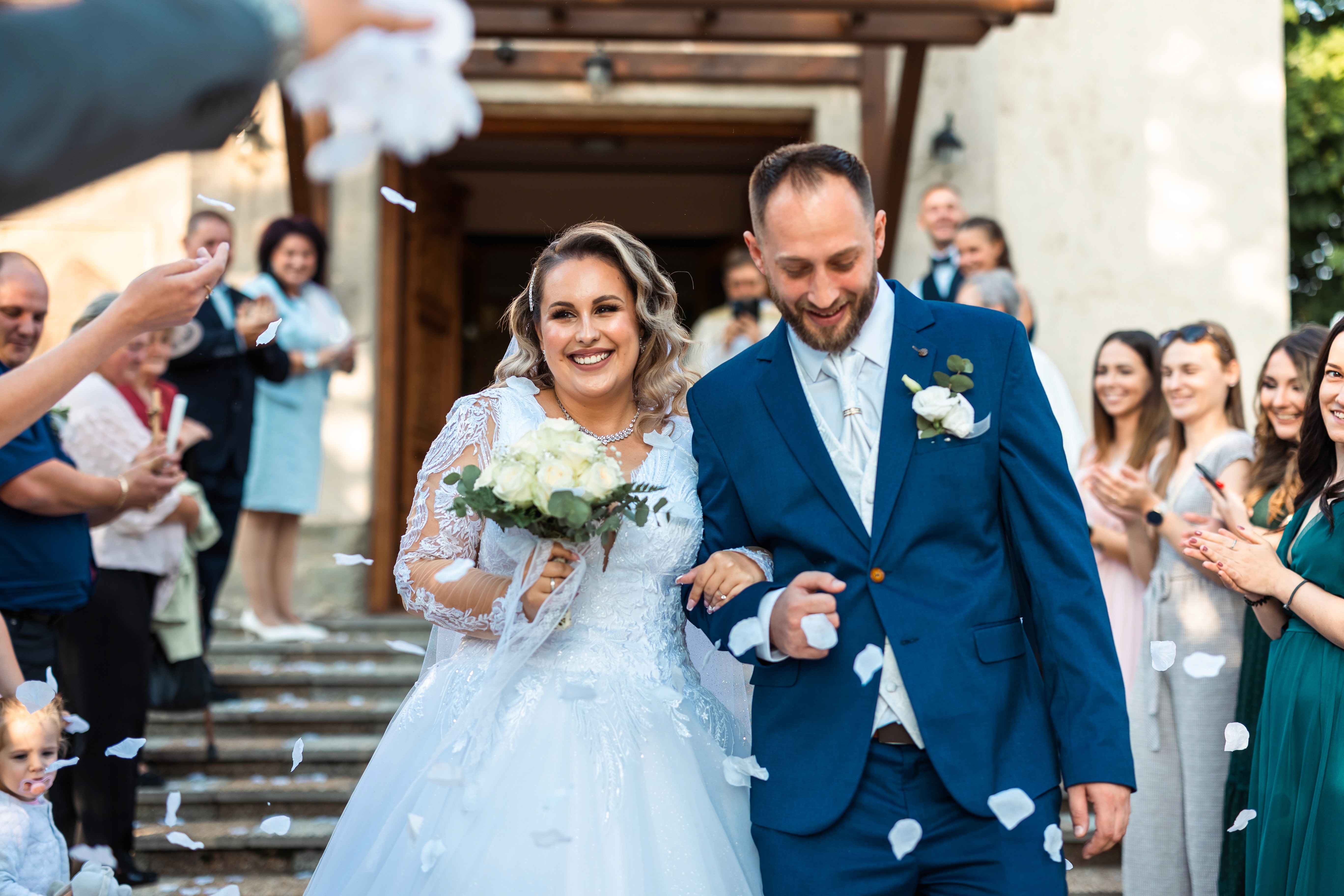 Mosolygó menyasszony csipkés ruhában fehér csokrot tartva és vőlegény kék öltönyben boldogan kilépnek a templomból sziromeső közepette, vendégek tapsolnak körülöttük