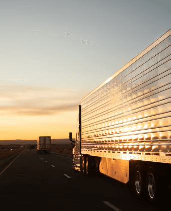 Refrigerated semi-truck trailer traveling on a highway at sunset, reflecting golden light off its polished aluminum sides.