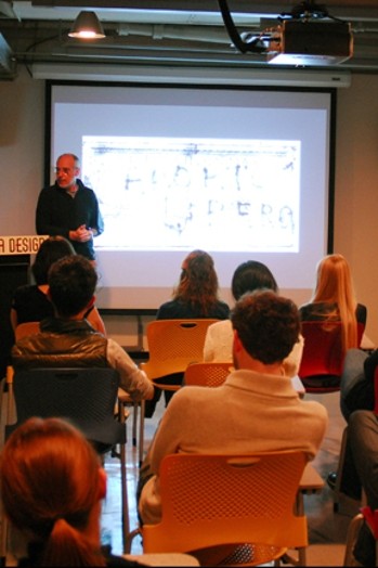 A man giving a lecture to a group of students while having behind him a screen projection.
