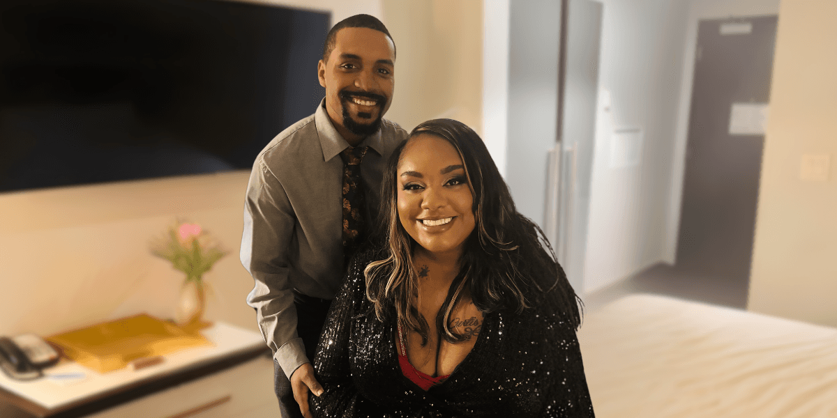 A smiling couple poses in a warmly lit room, exuding joy. The man wears a gray shirt and tie, and the woman wears a shimmering black top.