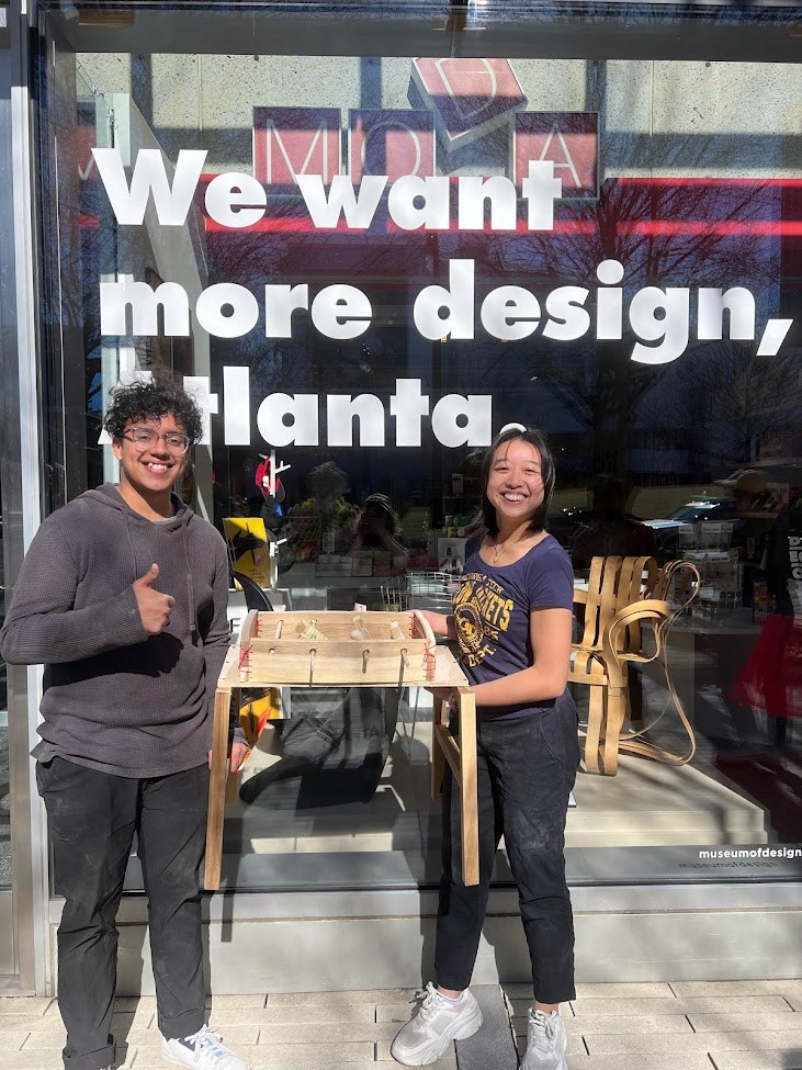 Woman sketching in a notebook at a desk with a plant backdrop.
