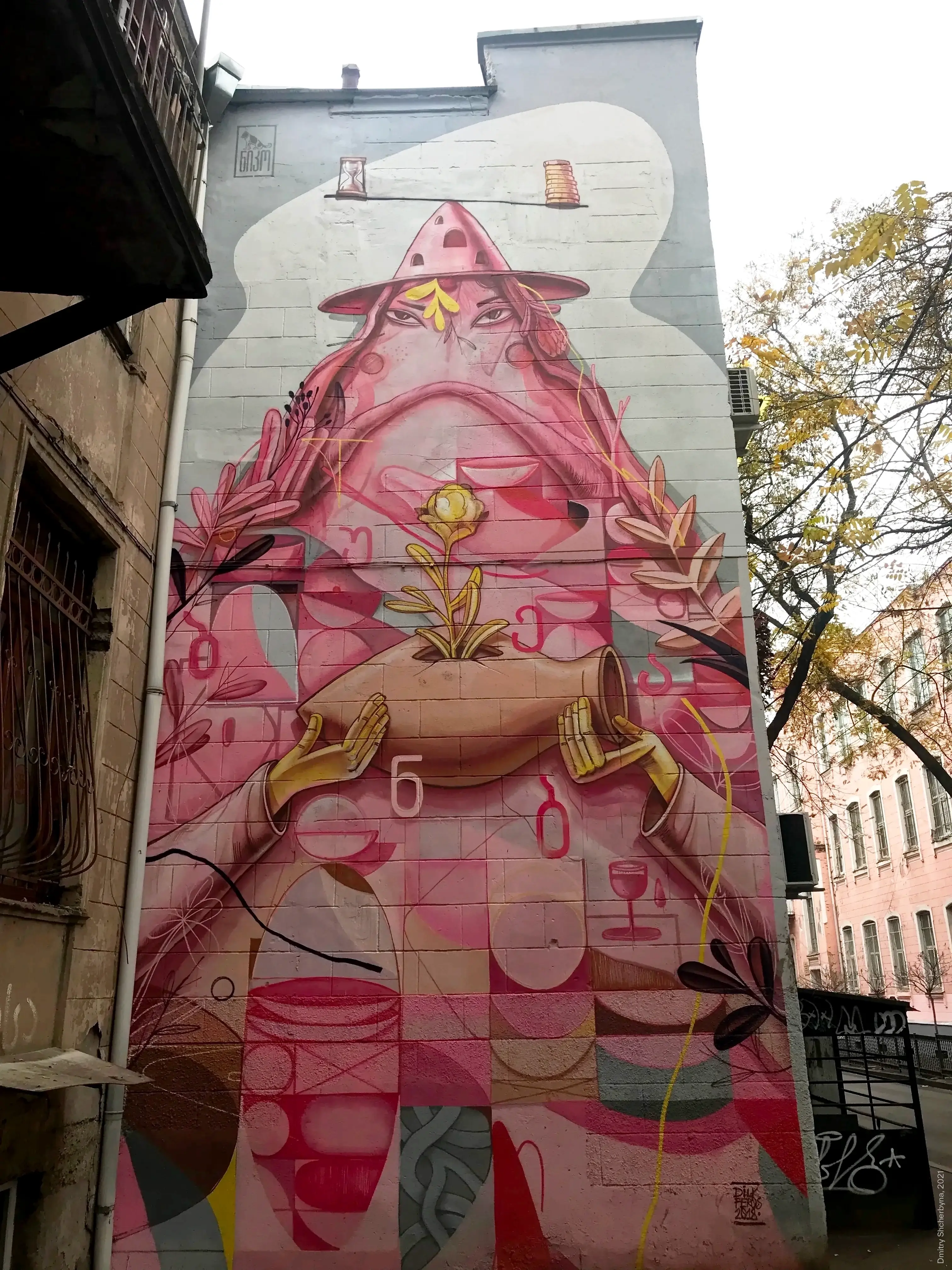 A large pink-toned mural depicting a stout figure wearing a pointed hat and holding a clay vessel containing a sprouting golden plant. Georgian alphabet letters and symbols related to wine are scattered throughout the composition.