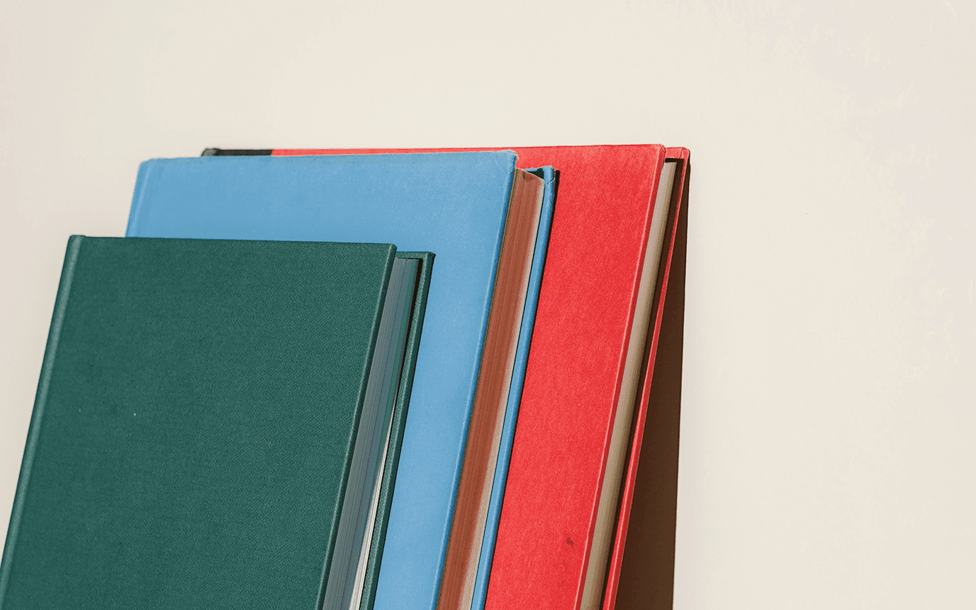A close-up of a stack of books in green, blue, red, and brown against a light background.