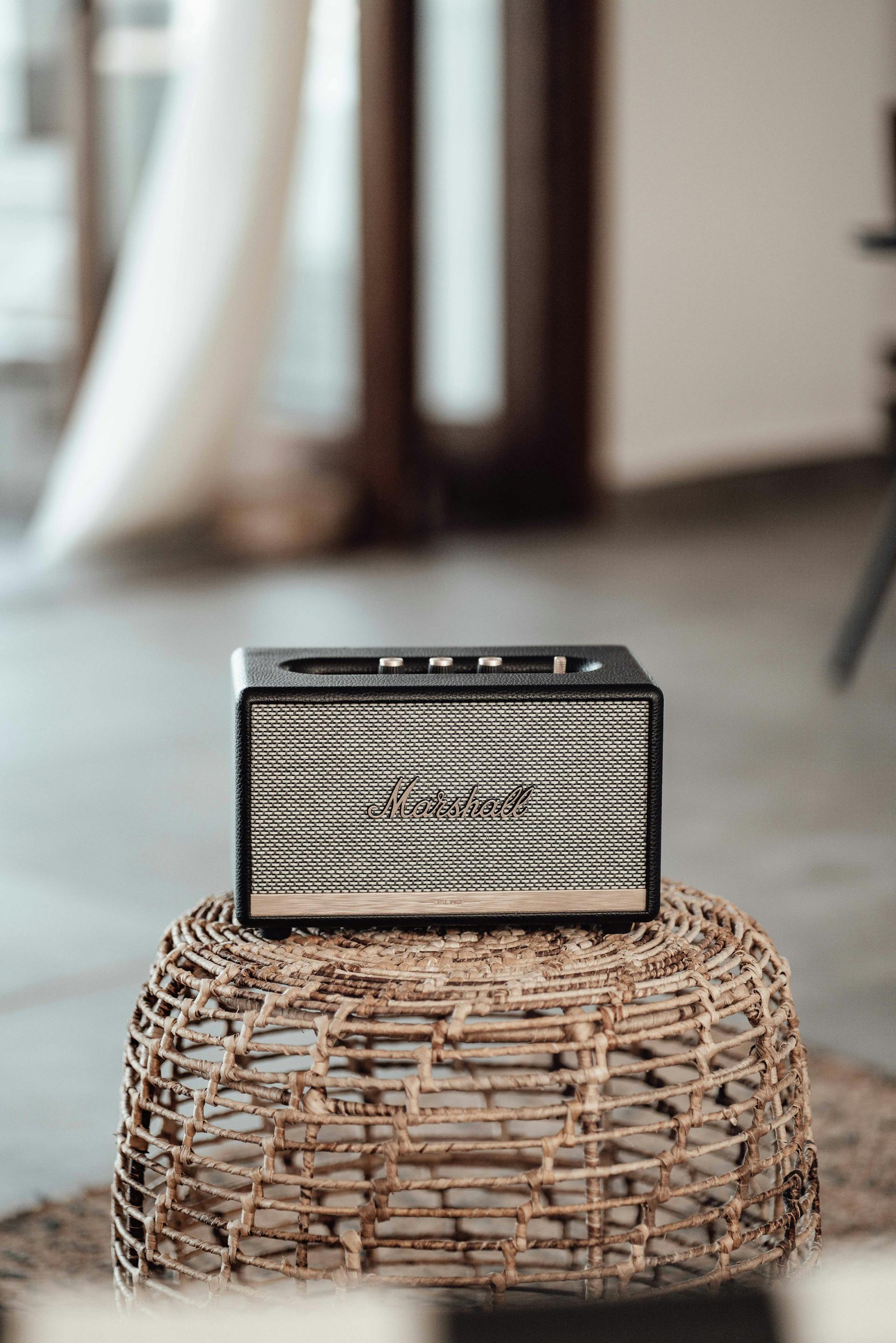 Black and tan Marshall Bluetooth speaker sitting on a woven rattan stool.