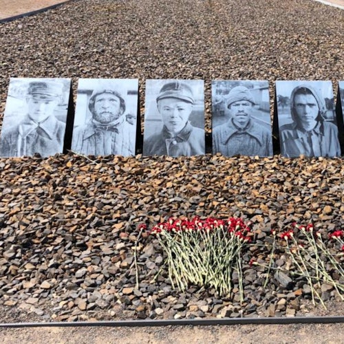 여섯 개의 흑백 초상화가 자갈 바닥 위에 나란히 놓여 있고, 그 앞에는 빨간 꽃이 놓여 있습니다.