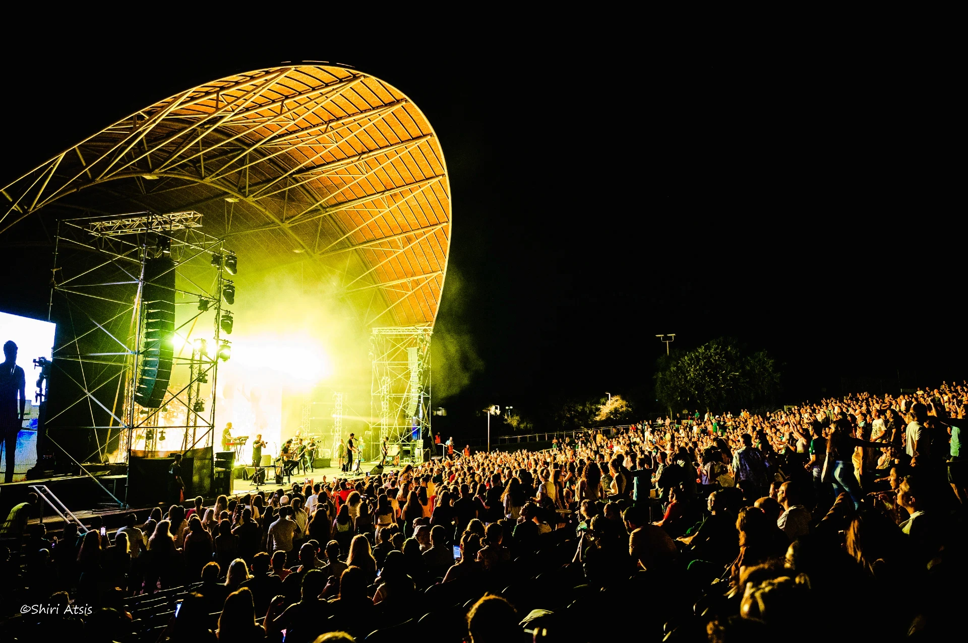מבט פנורמי מרהיב על הופעה חיה של עידן עמדי תחת כיפת השמיים. הקהל הרב ממלא את האמפיתיאטרון מול במה מוארת בתאורה צהובה חמה ועוצמתית מתחת למבנה קשת ייחודי, מאת Shiriz Photography הצלמת שירי עציץ.