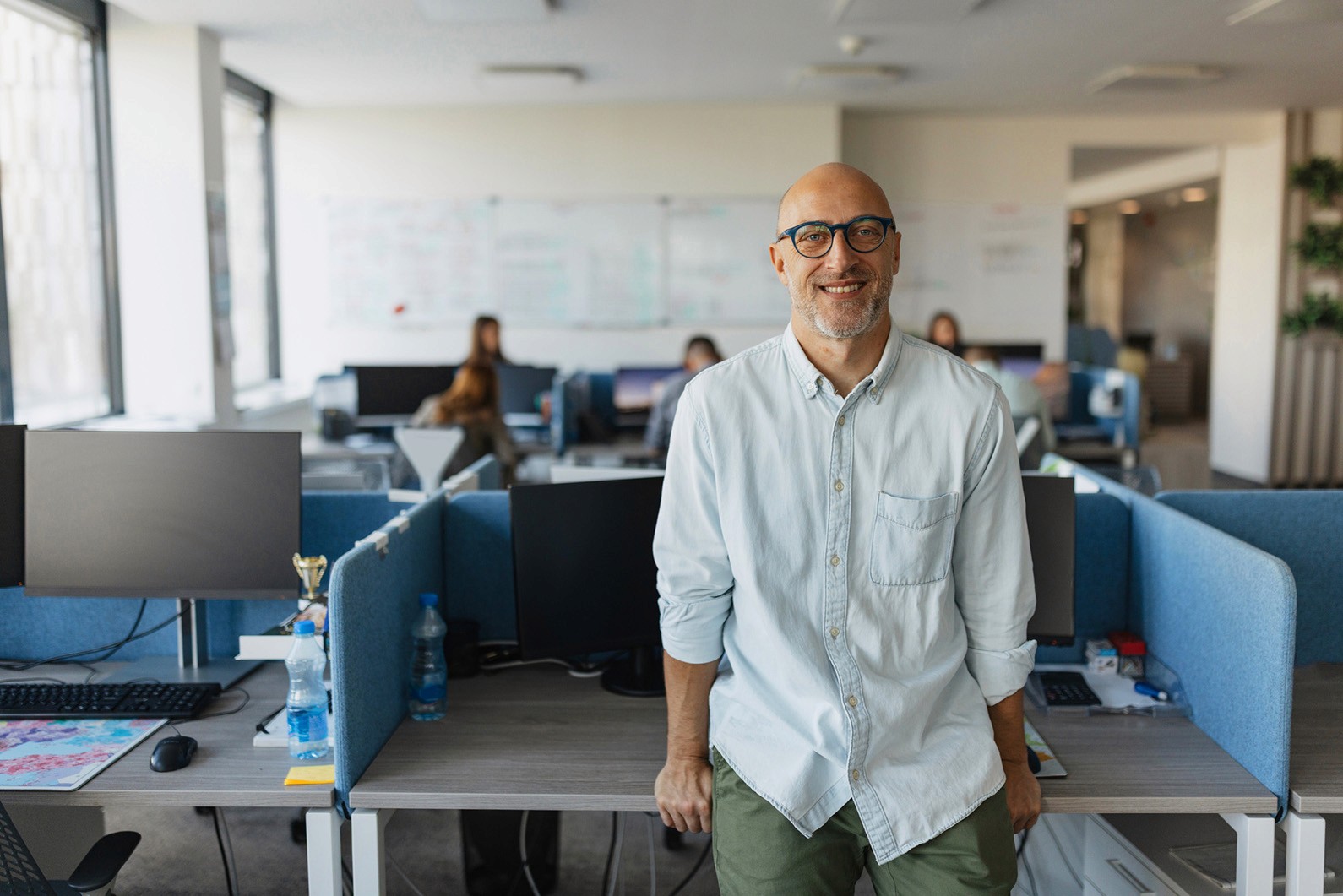 Happy bald man with glasses in a modern office, smiling at the camera, leaning on a desk with computers.