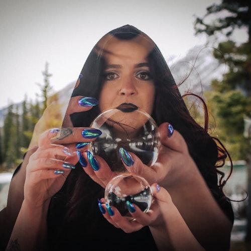 A woman with long dark hair and blue metallic nails holds two reflective spheres, with trees and mountains in the background.