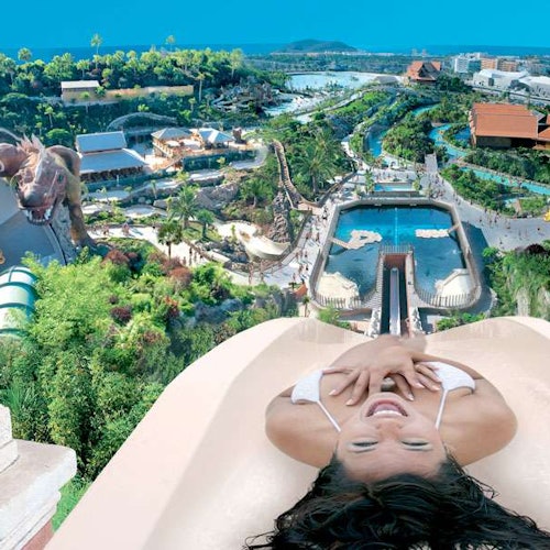 A woman sliding down a water slide, exhilarated, with a view of a large water park featuring pools, greenery, and attractions.