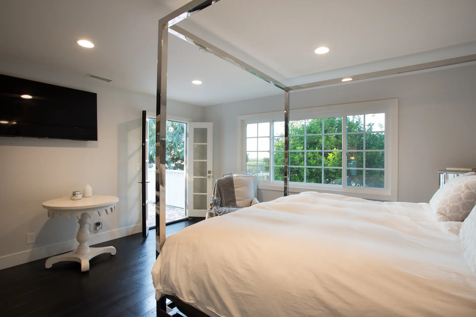 Elegant primary bedroom with modern chrome bedframe, dark hardwood flooring and wall-mounted TV. From Dana Point Cape Cod remodel, photo by Chris Darnall.