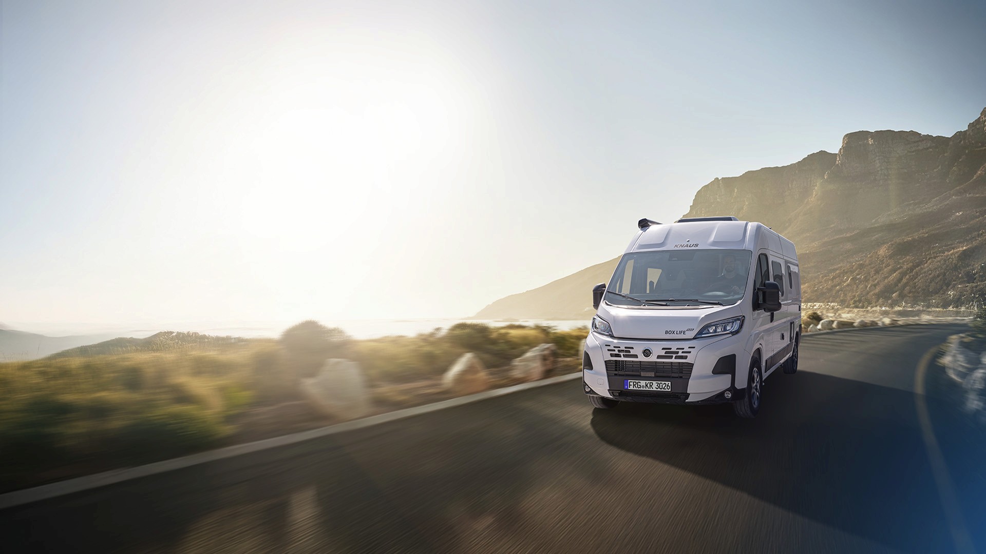 Das Bild zeigt einen KNAUS BOXLIFE PLATINUM CamperVan, der bei Sonnenaufgang eine kurvige Bergstraße entlangfährt.