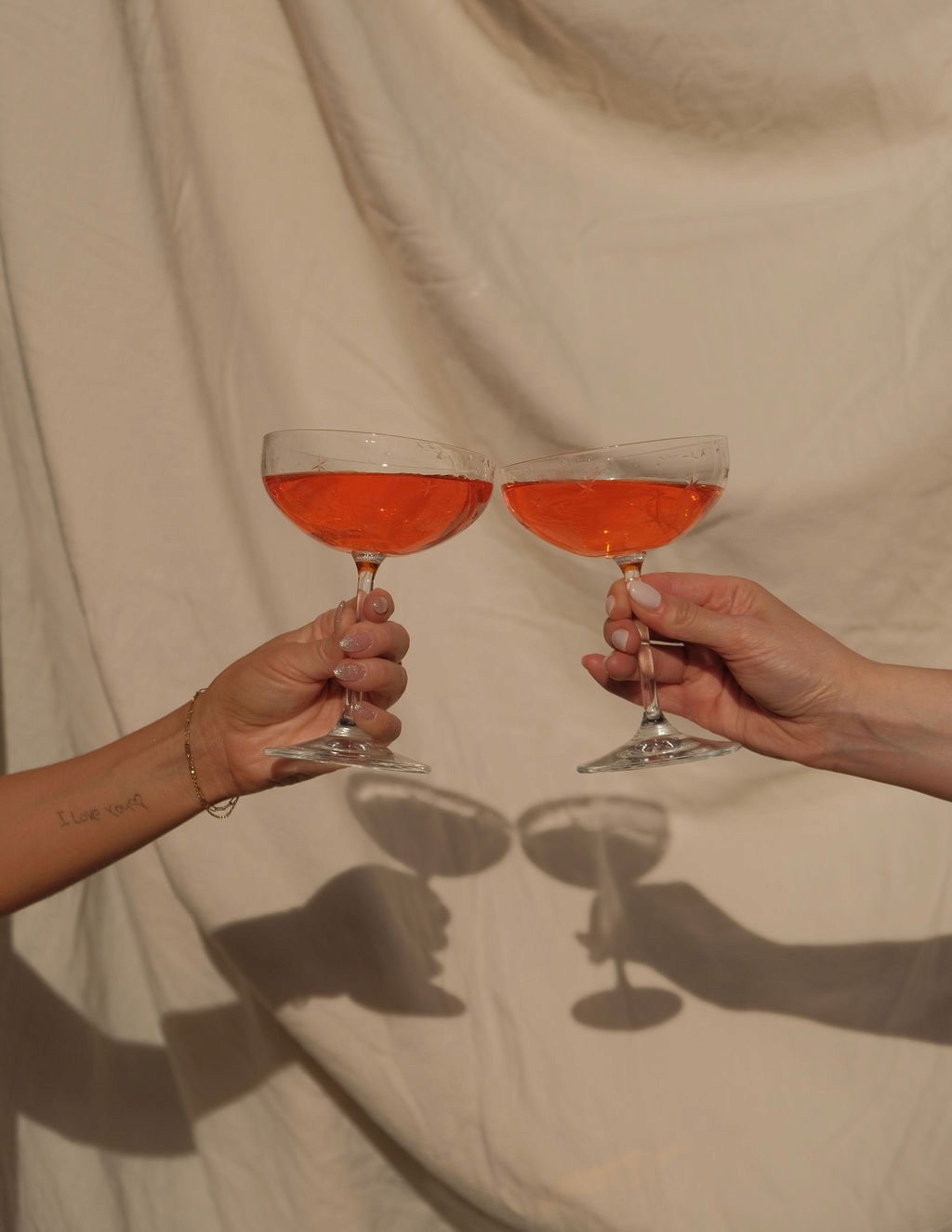 Duas mãos erguem taças de coquetel cheias de uma bebida vermelha ou rosé, fazendo um brinde. A cena é contra um fundo de tecido bege claro e amassado, com sombras projetadas das mãos e taças.