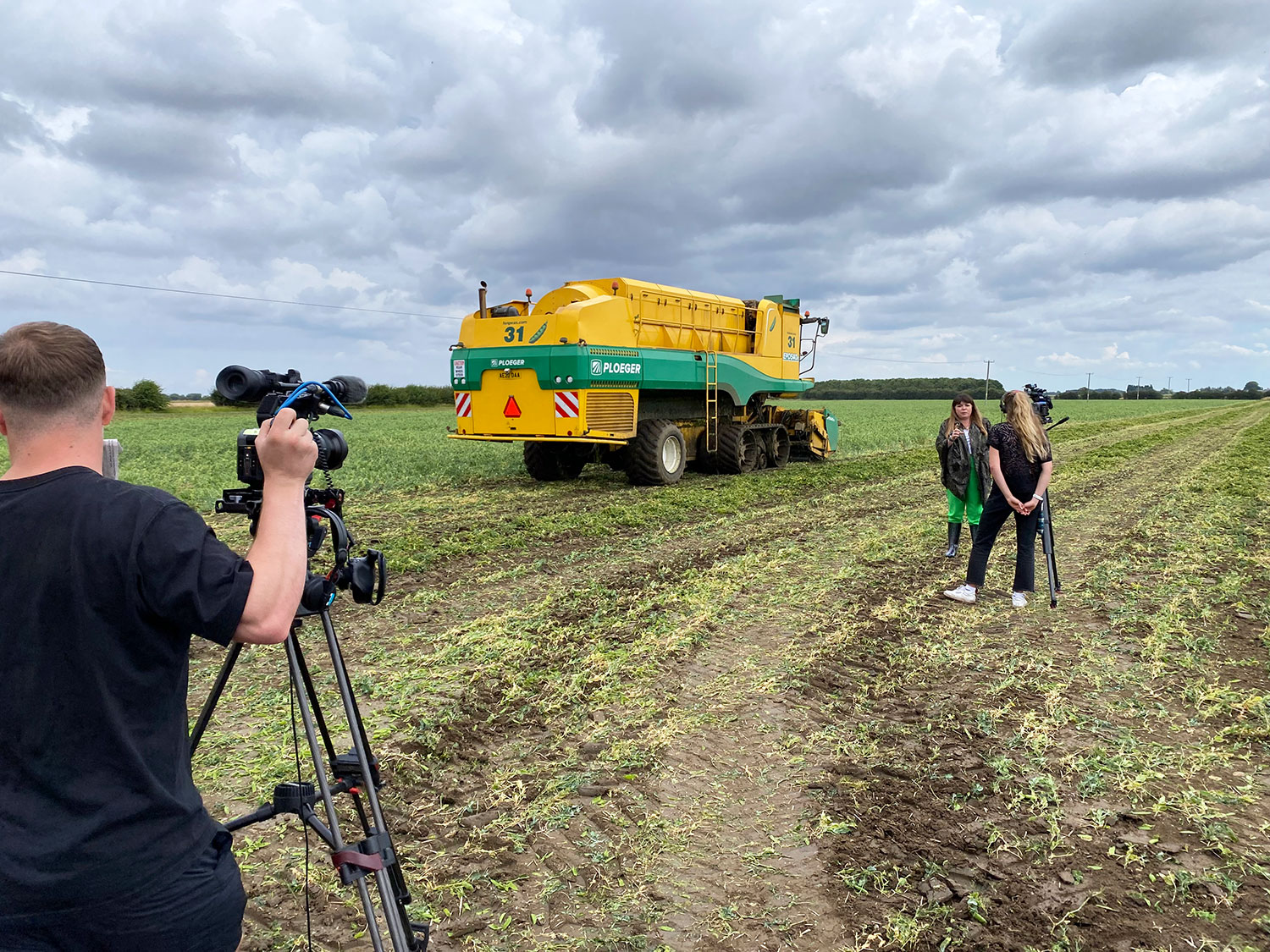 Briony Williams and two camera crew mewmbers standing in a field. Behind them is a yellow and green Ploeger pea harvester