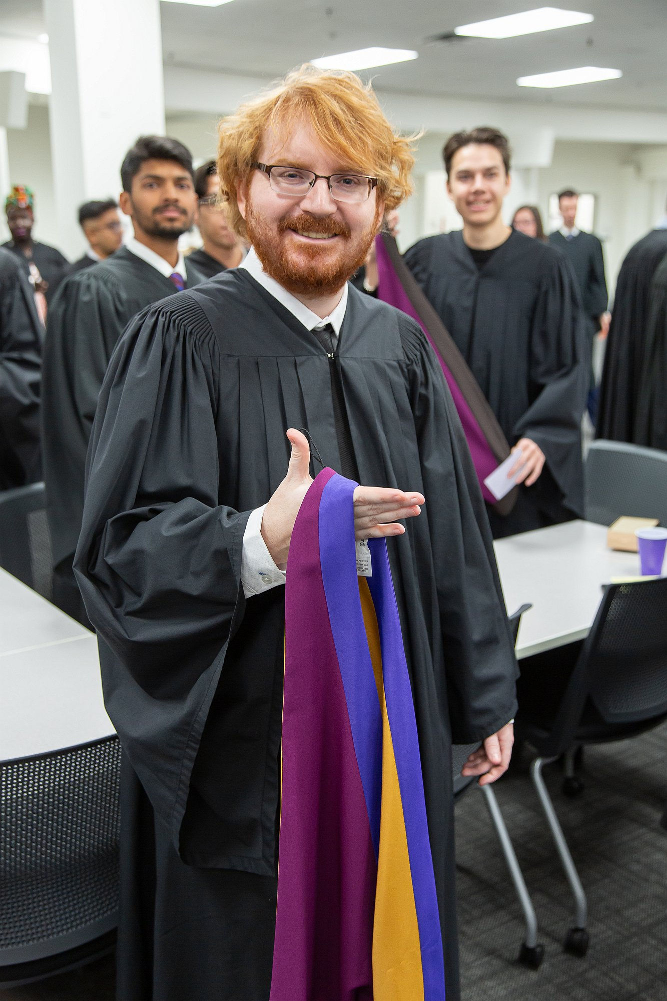I'm wearing a black graduation gown and smiling while holding colorful sashes in purple, yellow, and blue. I'm standing in a room with fellow graduates, and the energy feels celebratory as we get ready for the ceremony.
