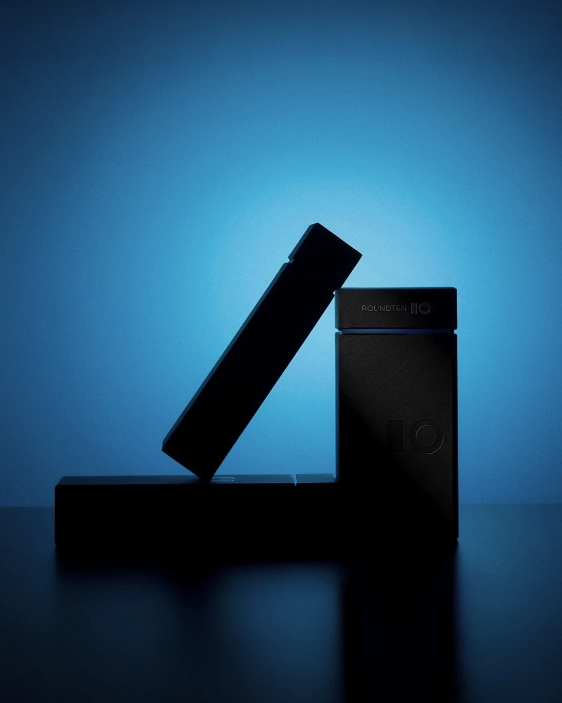 Black Roundten box leaning against a totem-shaped box in blue light, packaging design by Sebastian Saavedra