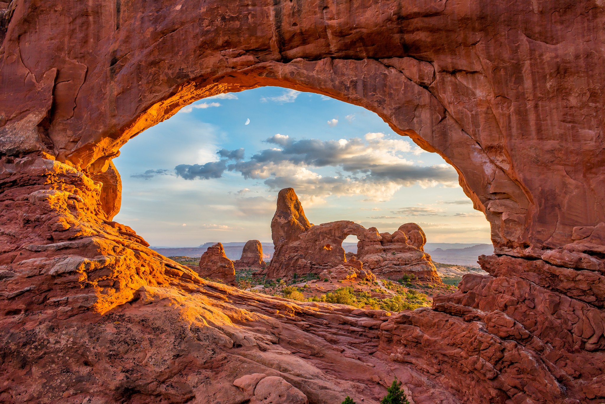 Sunlight illuminates a sandstone arch in Arches National Park, framing red rock formations and sky.