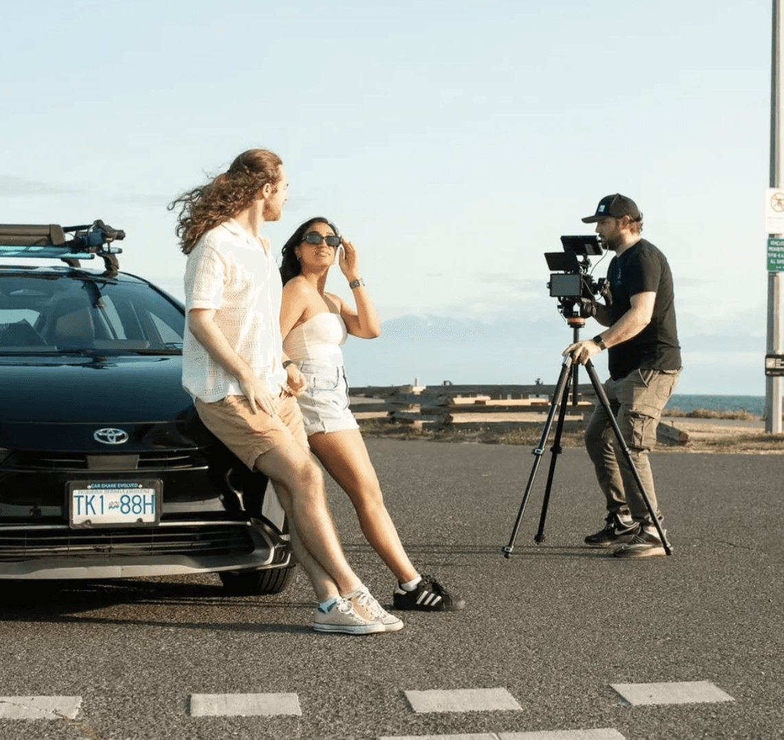 Two people by car look at camera, videographer filming