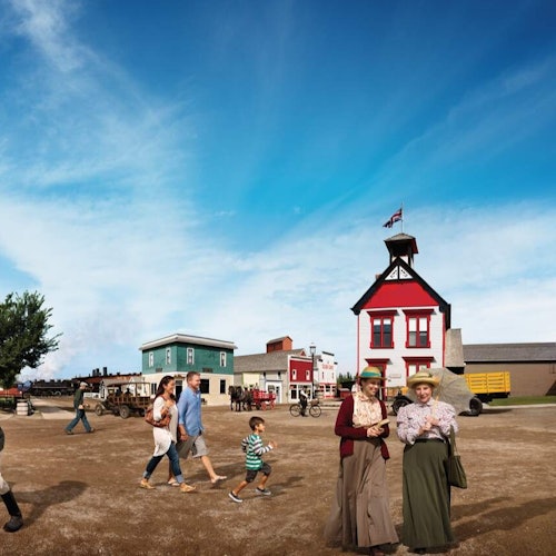 People walking in a historic village setting with old-fashioned buildings, including a red structure with a bell tower and flag.