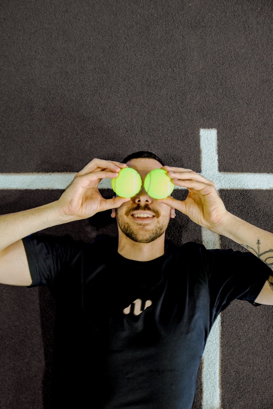 Padelspieler liegt lachend auf dem Court und hält zwei gelbe Bälle vor die Augen – ein verspielter Moment, der die humorvolle Seite von Padelnero zeigt.