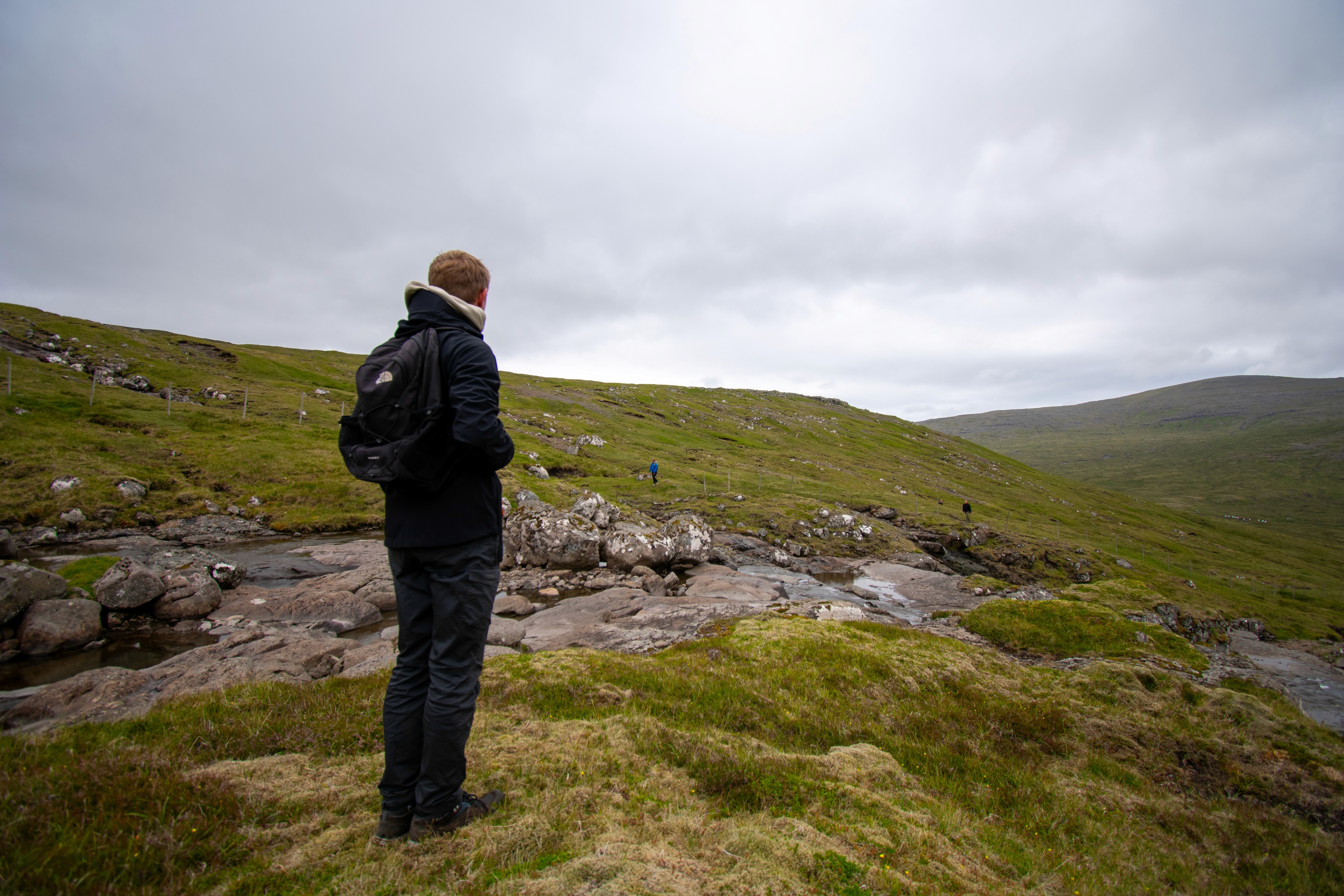 Silhouette of a person with a camera walking towards vibrant orange light under dim blue streetlights on a misty road at night. Mysterious and atmospheric.