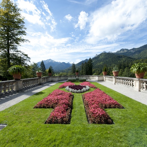 Upravená zahrada se vzorovanými květinovými záhony, obklopená kamenným zábradlím, s výhledem na hory a lesy pod částečně zataženou oblohou.