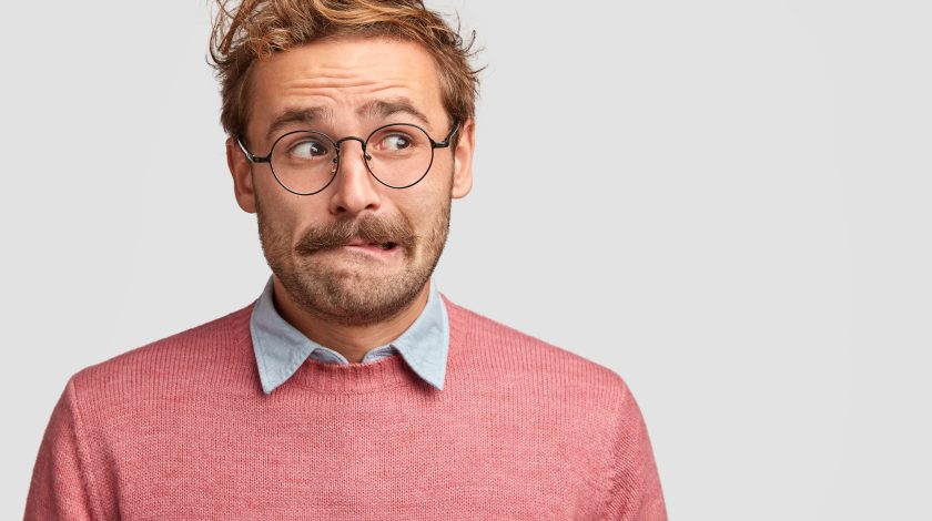 A man with a mustache and glasses looks to the side with a concerned expression, wearing a pink sweater over a blue collared shirt, against a plain gray background.