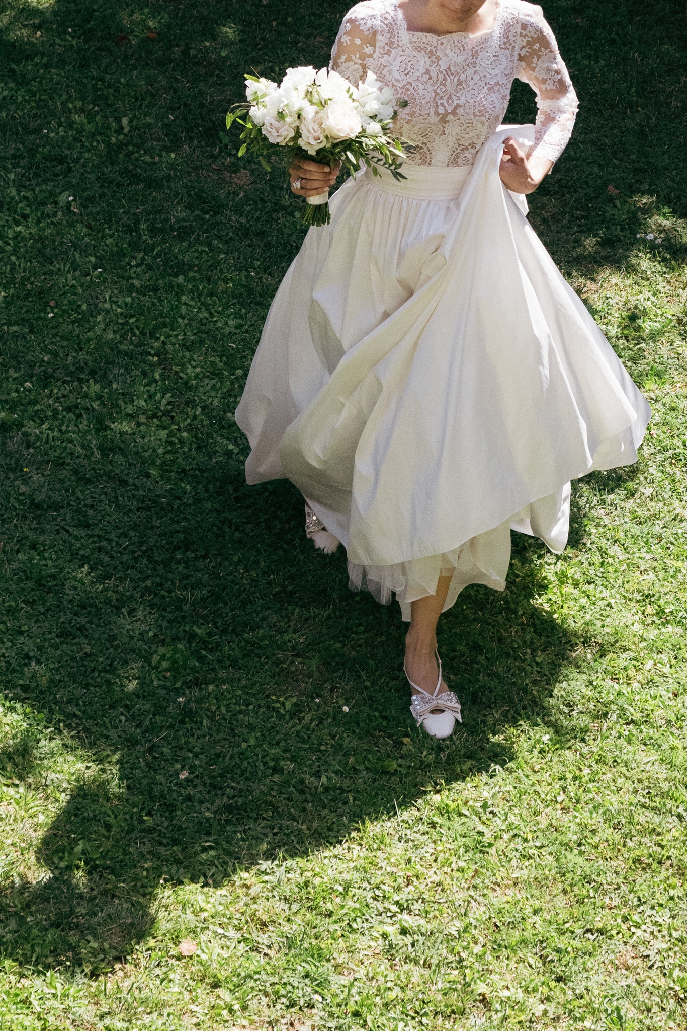 Sposa in abito bianco con corpetto in pizzo cammina sul prato tenendo bouquet di rose bianche, reportage di matrimonio