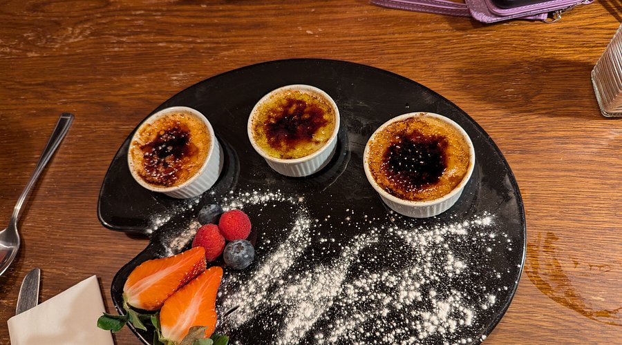 Gourmet dessert presentation featuring three caramelized custard tarts on black slate with fresh fruit garnish
