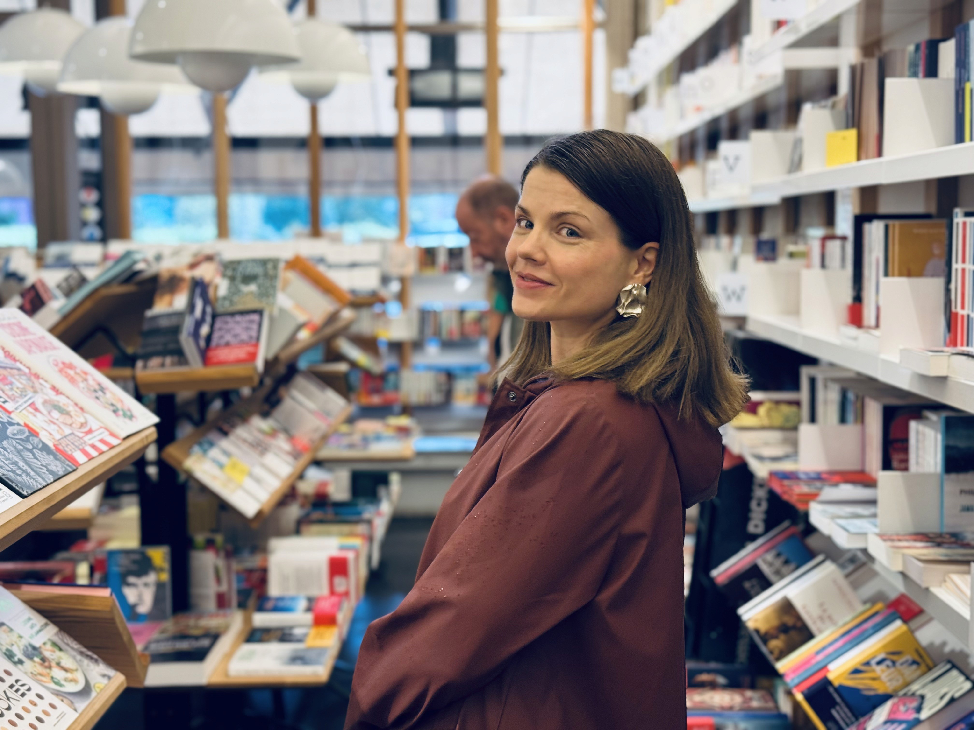 Tosca de Jong in a bookstore, wearing a burgundy coat and silver statement earrings, looking back over her shoulder among shelves filled with books.