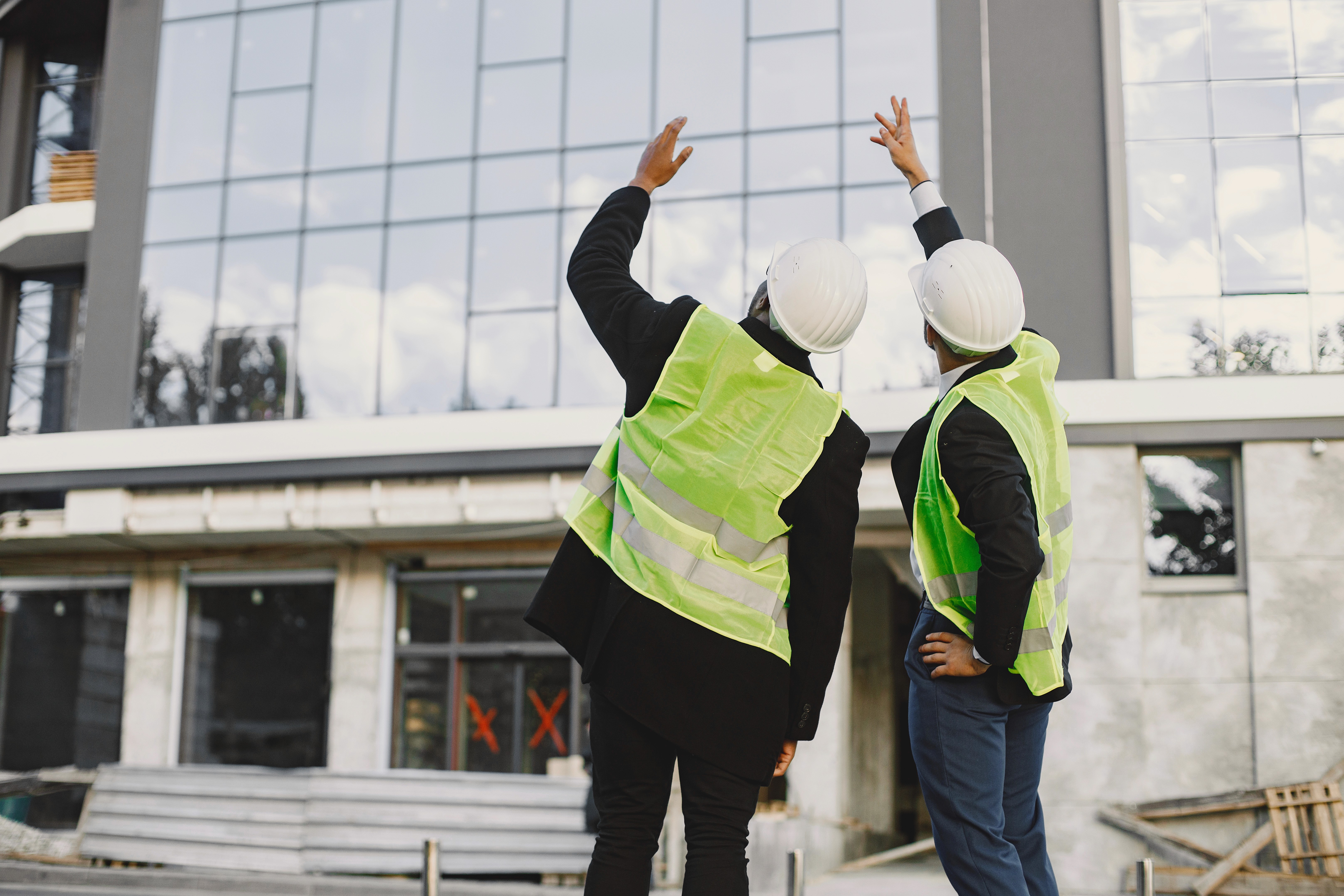 Two construction workers in safety vests and hard hats stand outside a modern glass commerical building, pointing upwards, conveying collaboration and focus.