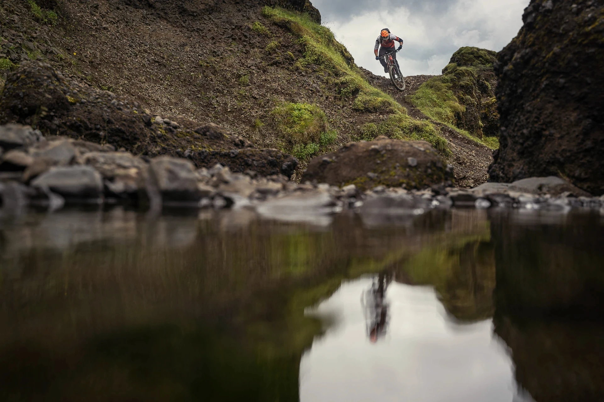 Mountain biker descending a rocky trail, reflected in a calm stream below.