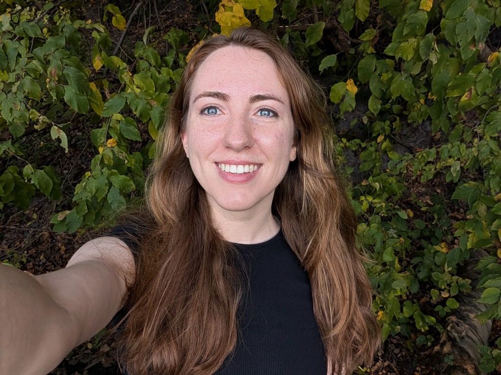 A photo of a woman with brown hair and blue eyes smiling at the camera with trees in the background