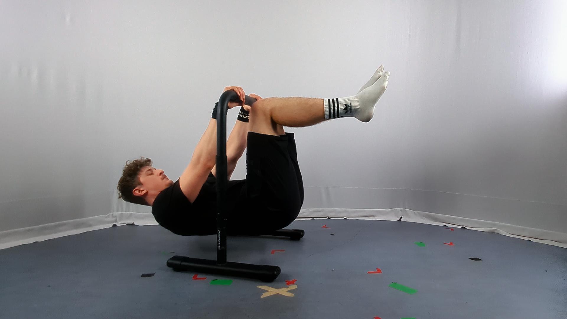 A person performs an advanced tuck front lever hold using parallel bars, demonstrating a calisthenics exercise technique in a minimalist indoor setting.