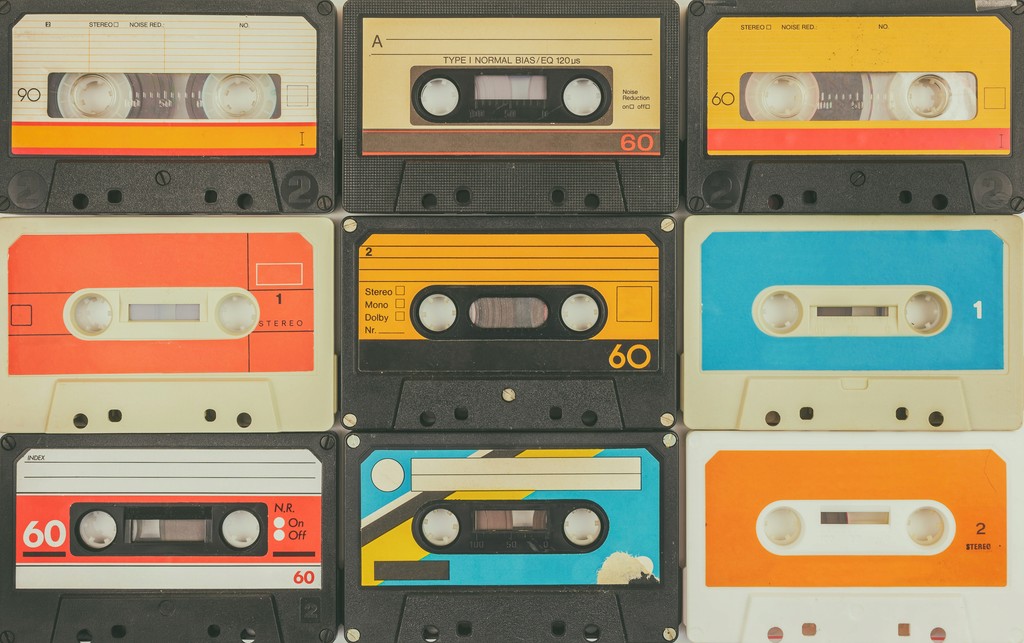 A collection of colorful vintage cassette tapes arranged in a grid pattern. The variety of designs and colors, including orange, blue, and yellow tapes, evokes nostalgia and highlights the retro charm of analog music media from the 1980s and 1990s.