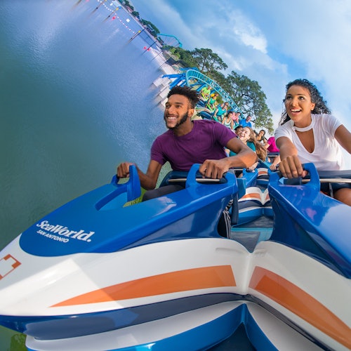 People smiling and enjoying a rollercoaster ride, with water and trees in the background.