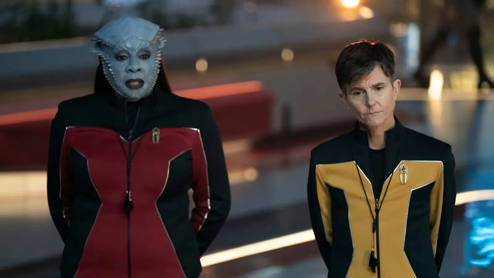 Two individuals in futuristic uniforms, one with alien features, stand in a brightly lit, modern setting with sleek architectural elements.