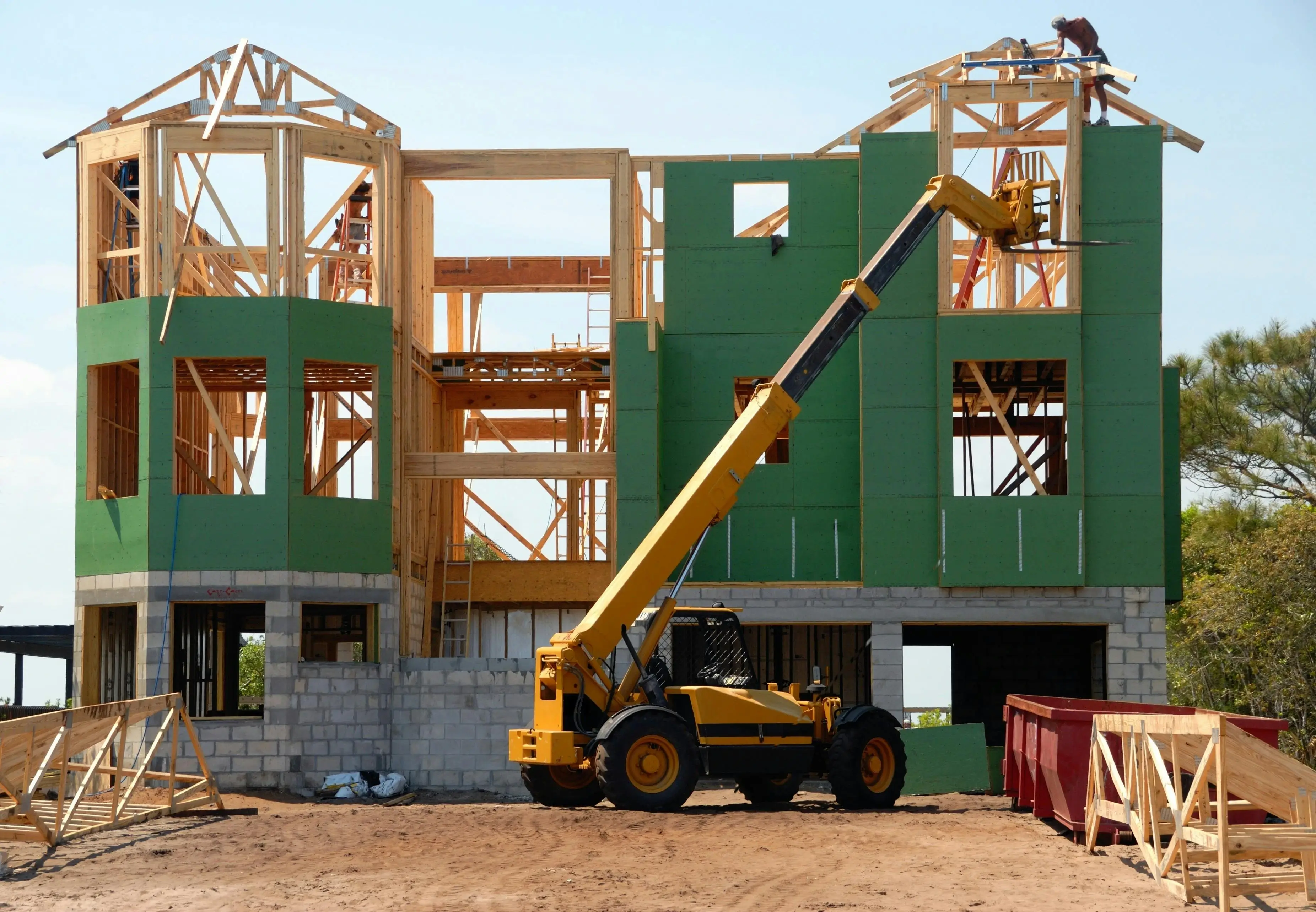 Construction site of a two-story house with exposed wooden framework and green sheathing. A yellow forklift hoists materials, and a worker stands on the roof.