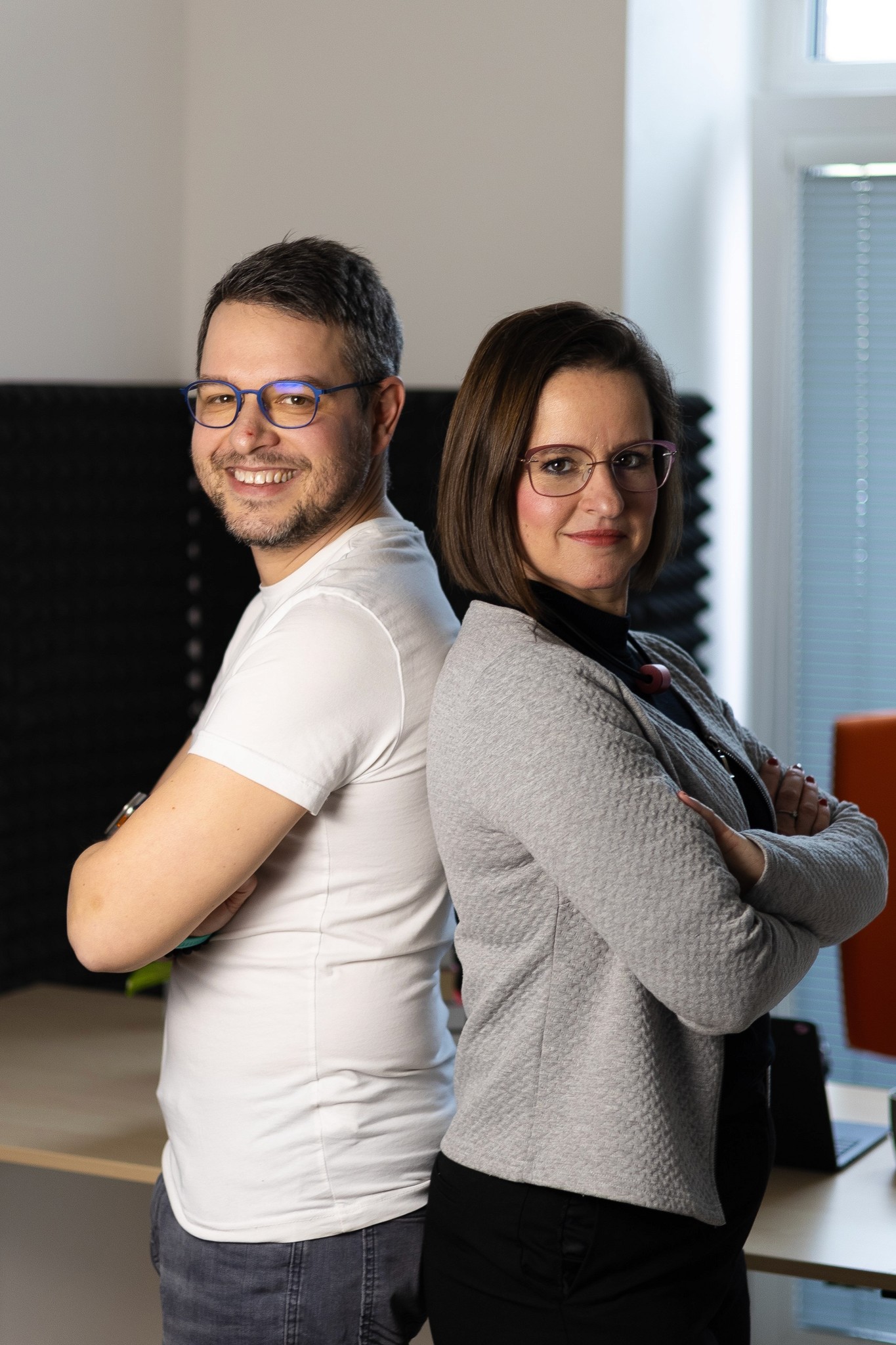 Dva usmívající se lidé stojí zády k sobě a vyzařují přátelské a sebevědomé vystupování v dobře osvětleném prostoru.