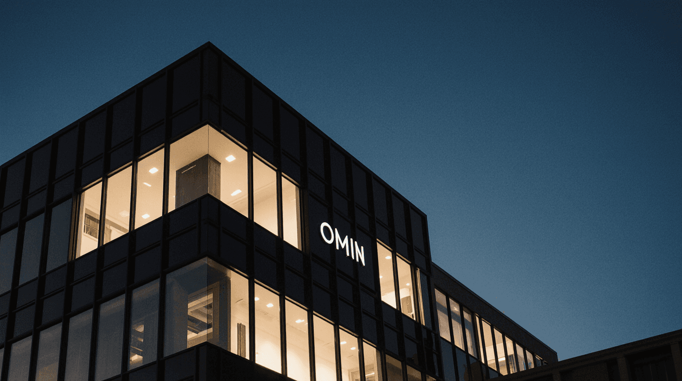 Modern glass office building at dusk with illuminated interiors and the word ‘OMIN’ displayed on its facade.