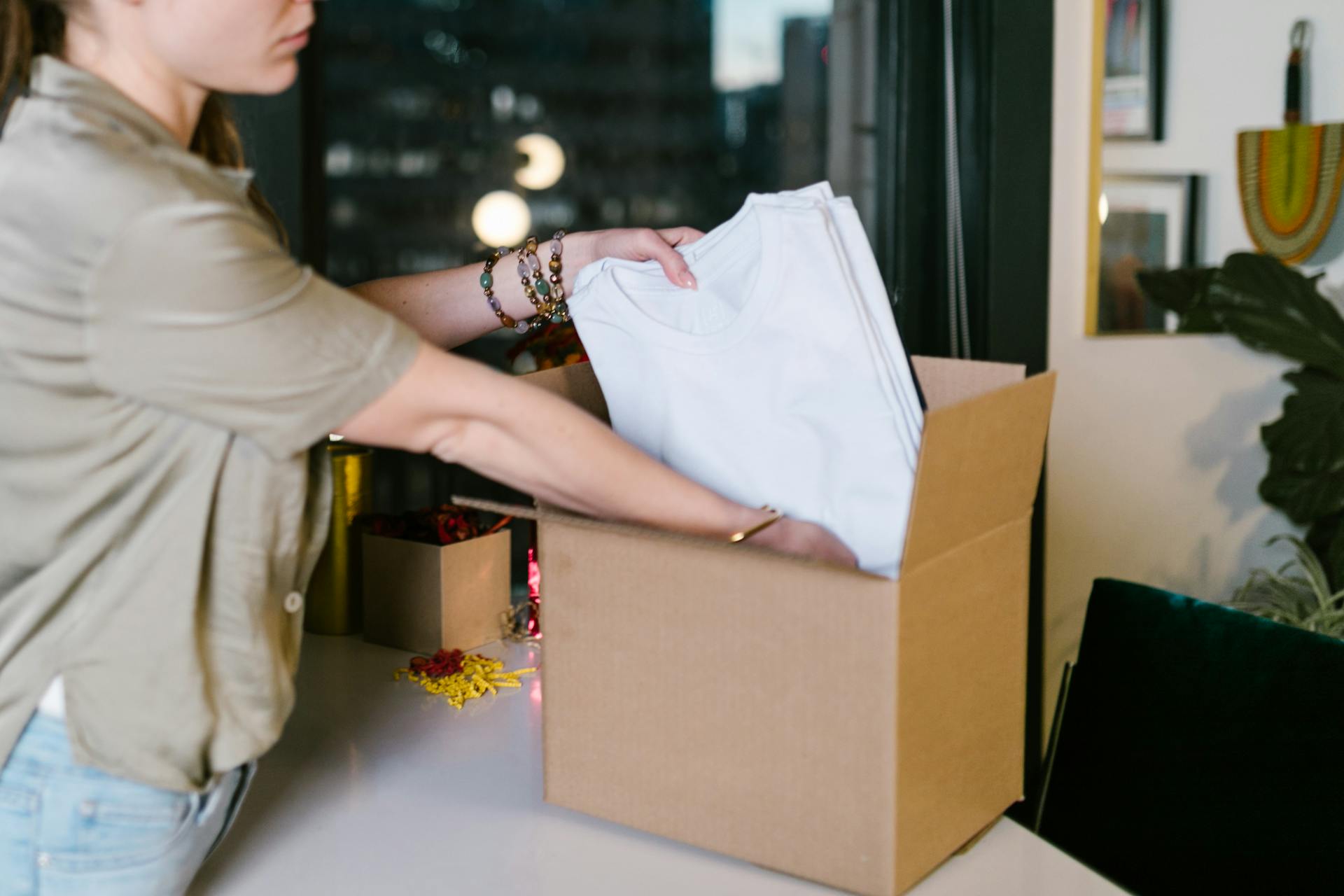 A person wearing a light green shirt and bracelets is placing a stack of neatly folded white t-shirts into an open, plain brown cardboard box. The background is a bit blurry but shows a window with a city night view and some wall art.