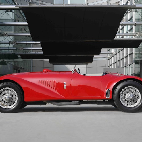 خودروی ورزشی کلاسیک قرمز کانورتیبل در یک محیط شهری مدرن با دیوارهای شیشه‌ای و پانل‌های بزرگ سیاه در بالای آن پارک شده است.