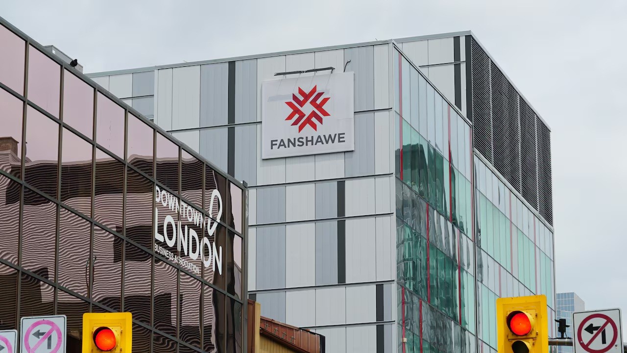 This image shows a modern building with a sign for Fanshawe College, a well-known post-secondary institution in London, Ontario, Canada. The building is located in the Downtown London area, as indicated by the reflection of another building labeled Downtown London Business Association. The architecture features reflective glass panels, and a red traffic light is visible in the foreground.
