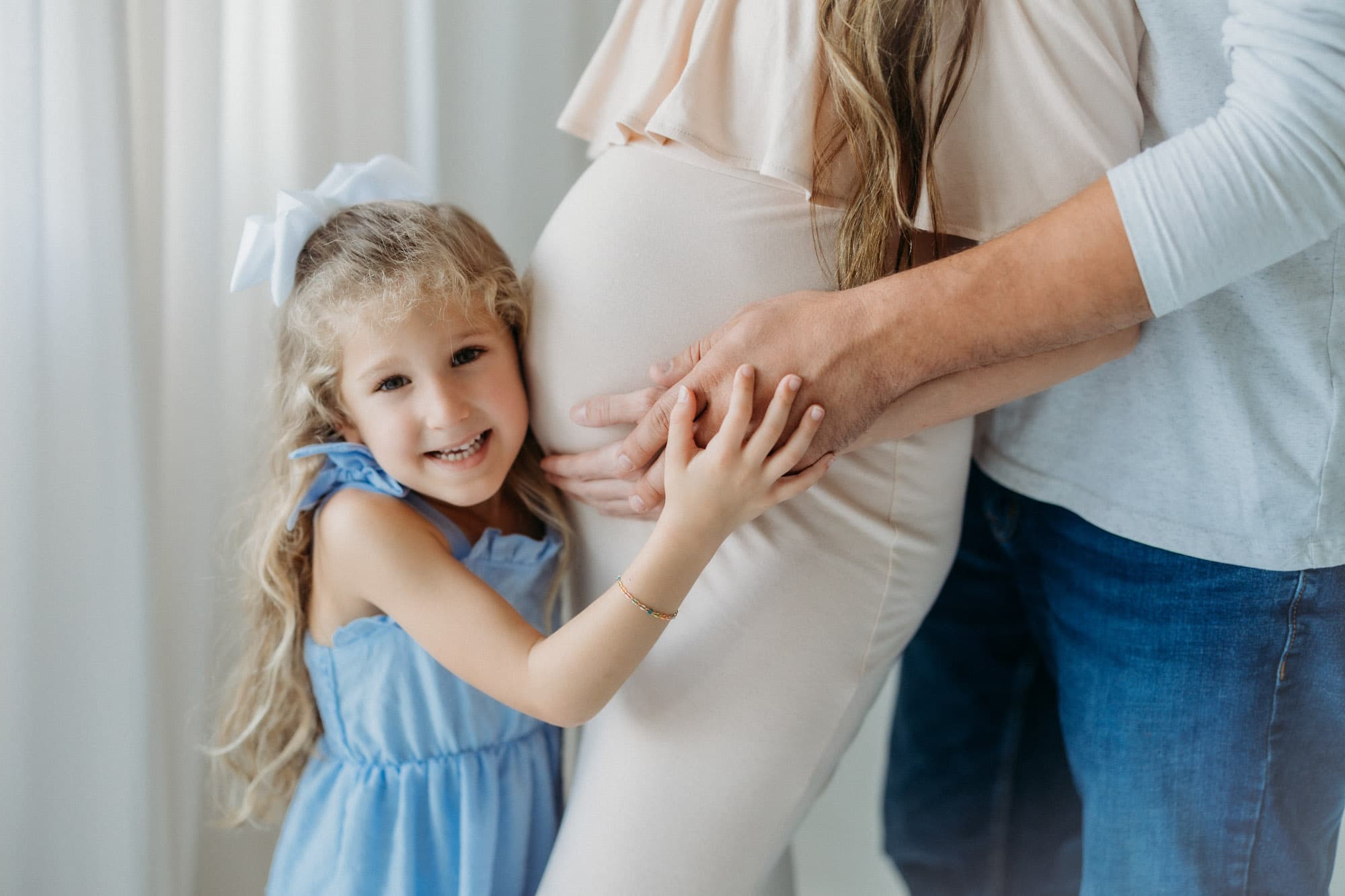 A young girl smiles while hugging her mother’s pregnant belly in the natural light of Revelator Studio, showcasing maternity photography in Shreveport.