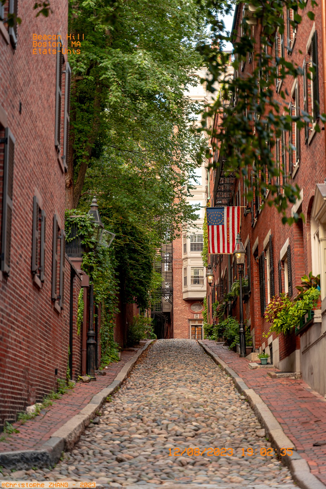 Rue pavée bordée de bâtiments en briques rouges à Beacon Hill, Boston, avec un drapeau américain.