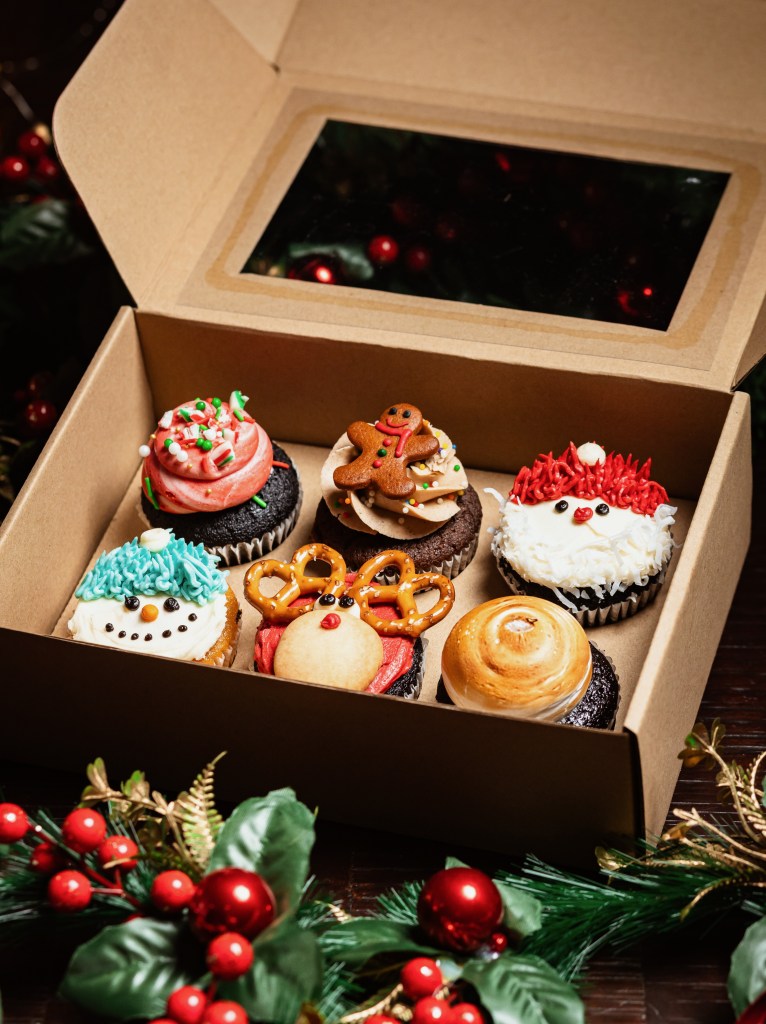 A festive box of beautifully decorated Christmas cupcakes, featuring designs like snowmen, reindeer, and Santa. The box is made of kraft paper and showcases a variety of colorful toppings, set against a backdrop of holiday decorations.