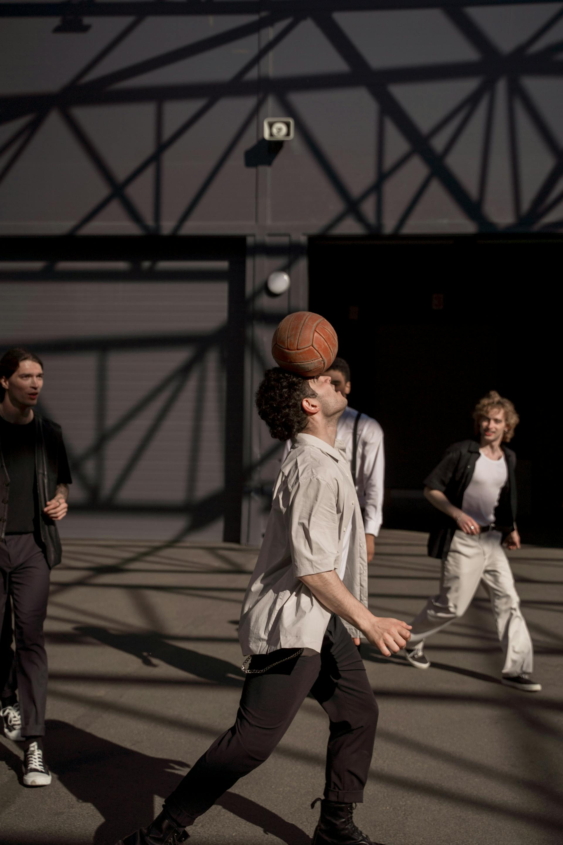 A man balances a vintage leather ball on his forehead while walking, with three other men watching him in an open outdoor area with strong shadows.