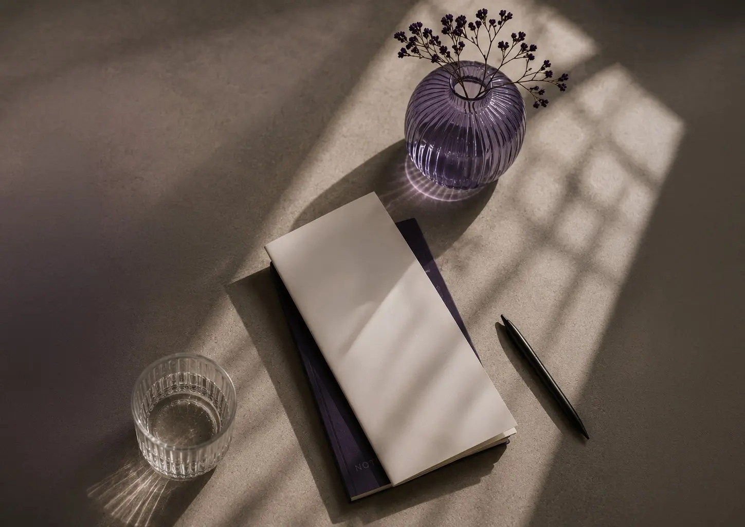 A laptop and a purple cup are placed on a wooden desk, casting shadows in the soft light.
